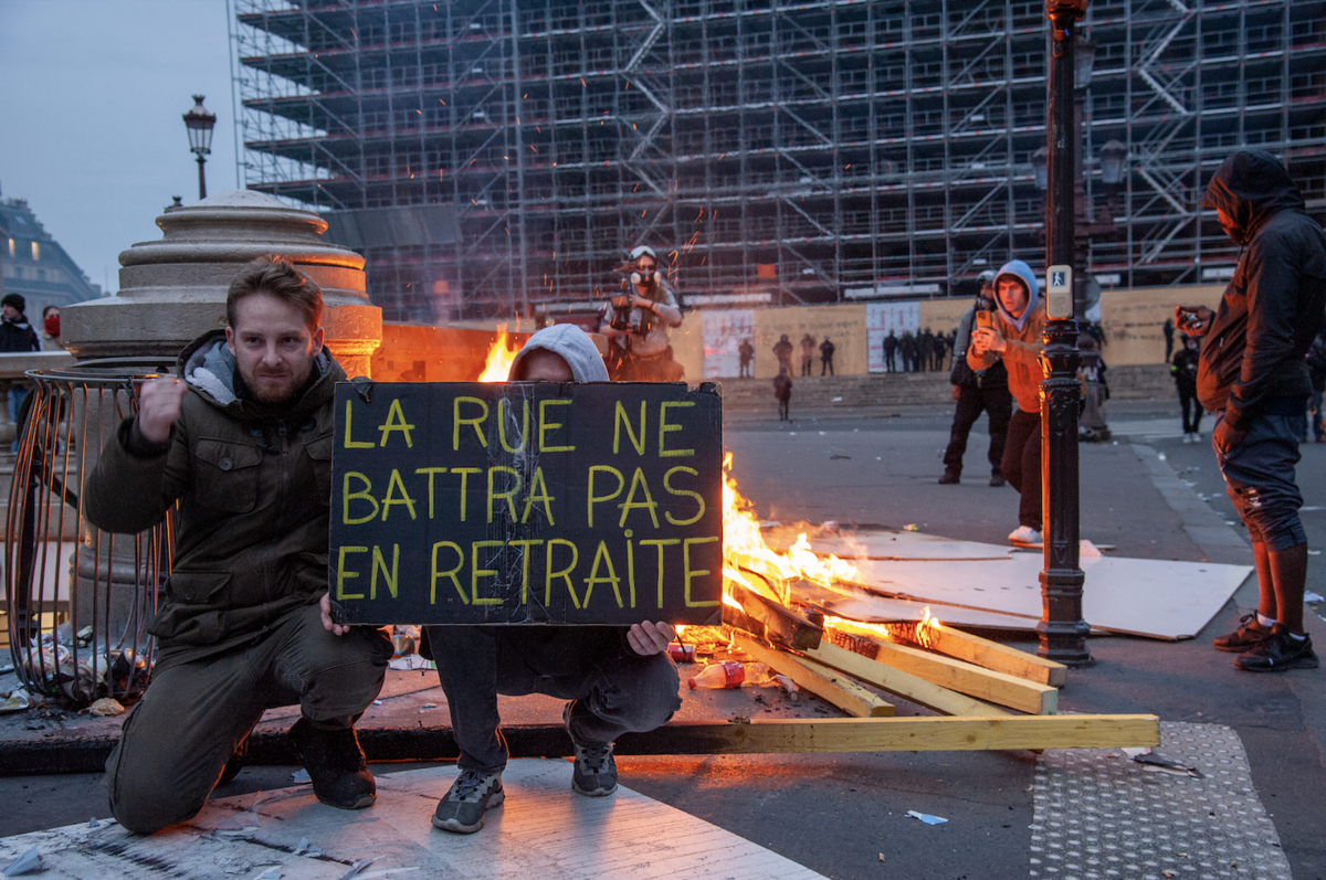 Deux manifestants prennent la pose devant un feu de palettes à côté de l'opéra avec une pancarte déjà croisée plus tôt. - © Reflets