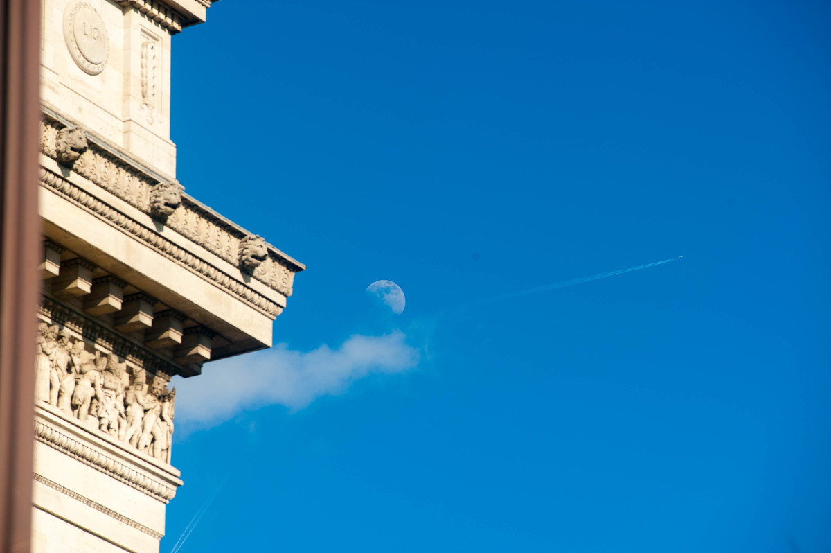 Au dessus de l'Arc de Triomphe, la lune regarde avec désintérêt ce qui se passe tandis qu'un avion traverse le ciel sans avoir la moindre idée de ce qui se passe au sol. - © Reflets